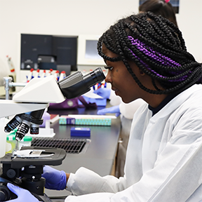 Medical Laboratory Technician student wearing white lab coat and gloves looking into a microscope.